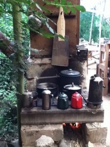 森の中にしつらえられた焚火の煮炊き場の上に並ぶ、赤や深緑やいぶし銀のポットや鍋などの道具